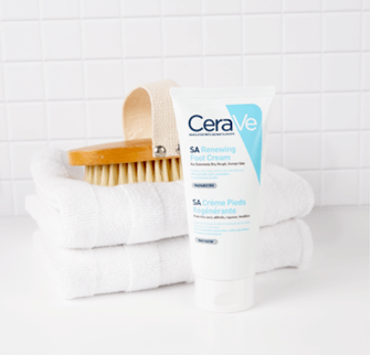 SA Renewing Foot Cream next to a foot exfoliating brush, which is sitting on two hand towels, against a white tiled wall